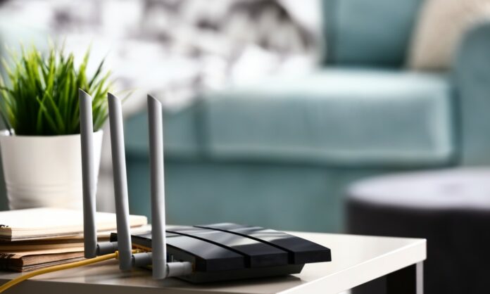 A black Wi-Fi router sitting on a coffee table in a home's brightly lit living room with a plant and a couch in the background.