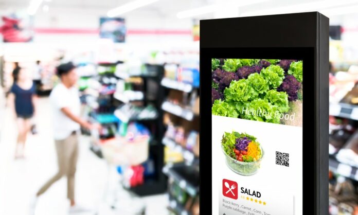 A digital signage stand advertising a salad in a supermarket with shoppers in the background.