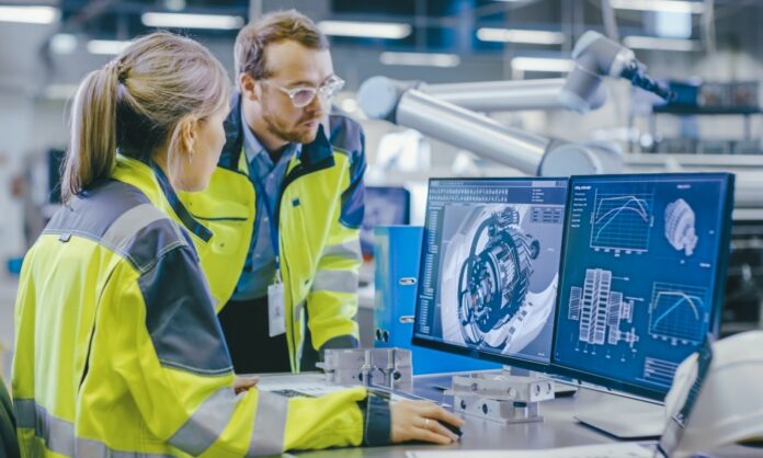 A female engineer examines digital data at the computer inside a manufacturing facility and communicates with a male colleague.