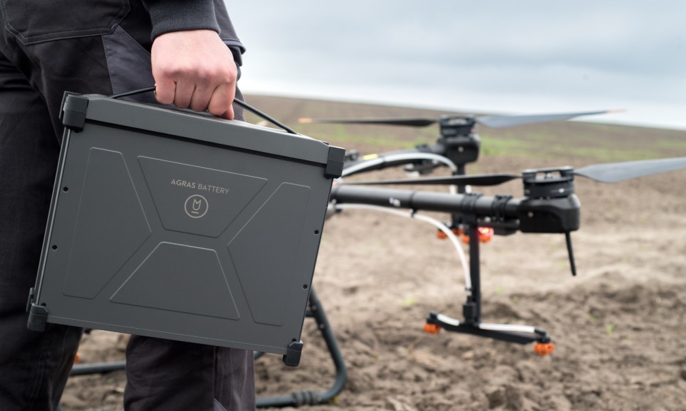 An unidentifiable man holding a black Agras battery case walking toward a drone sitting atop a patch of dirt.