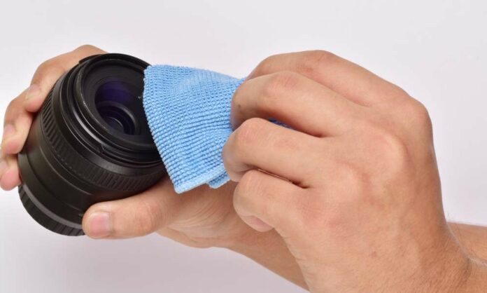 A close up view of a person cleaning a detached black camera lens with a small blue microfiber cloth.
