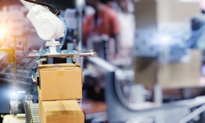 An industrial robot arm is packaging a box that’s rolling down a line on a factory floor in front of a blurred background.