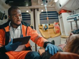 A man holds a tablet in one hand while resting his other on a patient’s shoulder in an ambulance.