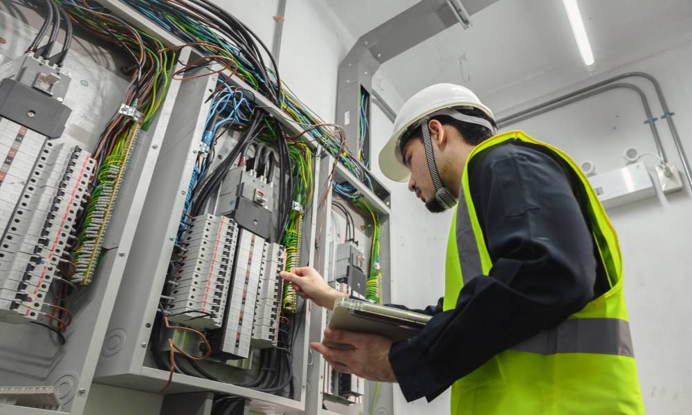 A man in a hard hat and neon vest inspects an electric panel in a small room while holding a clipboard.