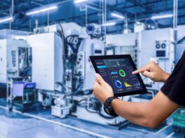 A person holding a tablet displaying charts and statistics stands in a brightly lit manufacturing facility.