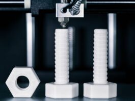 White 3D printed screws and bolts underneath a 3D printing machine. They stand on a flat, reflective surface.