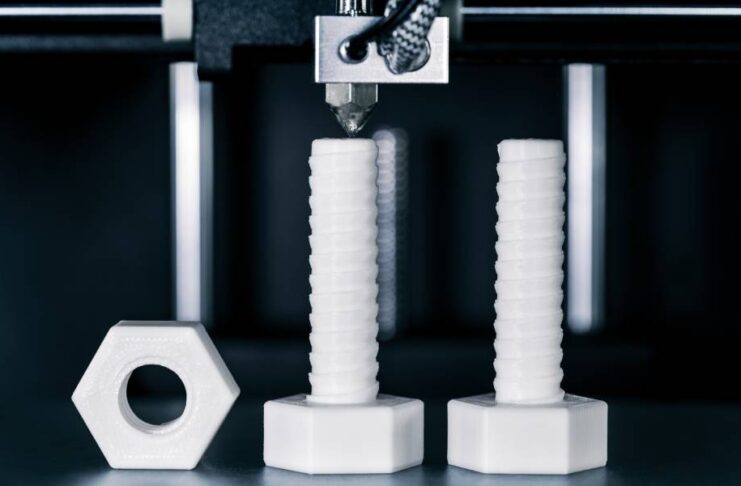 White 3D printed screws and bolts underneath a 3D printing machine. They stand on a flat, reflective surface.