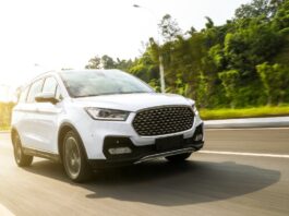 A white SUV drives down a rural road that is surrounded by green grass and tall, mature trees. The background is blurry.