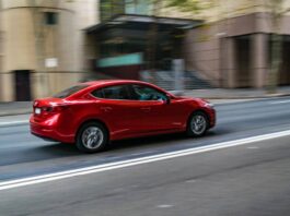 A red four-door sedan drives down an urban street. The building and tree in the background are blurry.