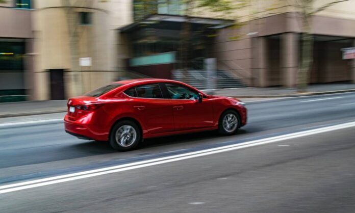 A red four-door sedan drives down an urban street. The building and tree in the background are blurry.