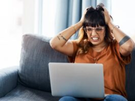 A woman grimacing and grabbing her hair in frustration as she sits on a couch and glares at the laptop in her lap.