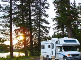 A white RV is parked in a secluded area surrounded by trees and water. Two tan lawn chairs sit in the grass next to the RV.