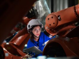 A female engineer wearing a blue jumpsuit and gray hardhat holding a clipboard inspecting a robotic arm in a warehouse.