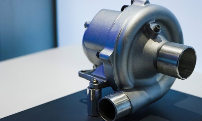 A close-up of an electric turbocharger with intake and exhaust pipes, displayed on a platform against a blurred background.