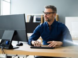 A businessperson sitting at his desk using a computer monitor and PC. He's wearing a navy button up and glasses.