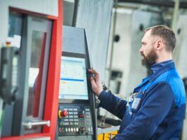 A bearded man in a blue jumpsuit works on the interior monitor of a machine in an industrial setting.