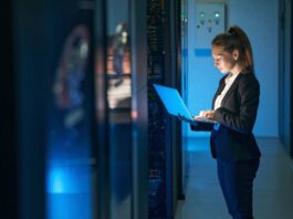 A woman in a black business suit with a ponytail stands in front of data centers holding and using a laptop.