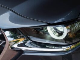 A close-up of a lit headlight on a grey-blue SUV with a round headlamp. The chrome insert reflects several trees.