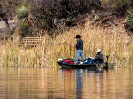 Two men in cowboy hats hold fishing poles while in a boat with a cooler and other supplies surrounding them.