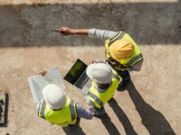 An overhead view of three construction workers wearing hard hats while holding blueprints and a laptop on a jobsite.