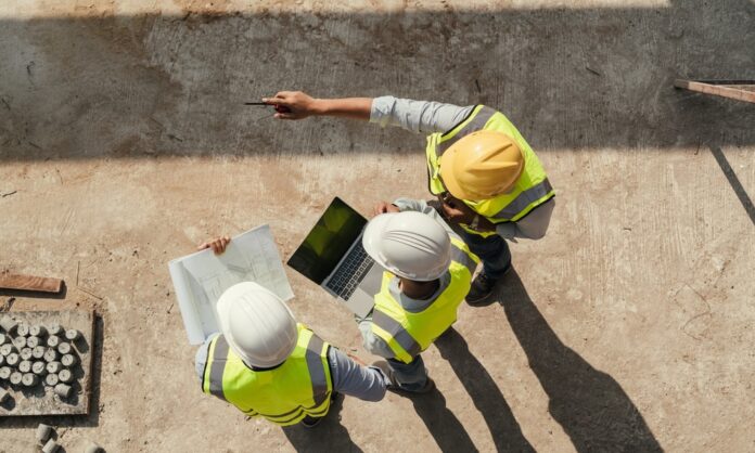 An overhead view of three construction workers wearing hard hats while holding blueprints and a laptop on a jobsite.