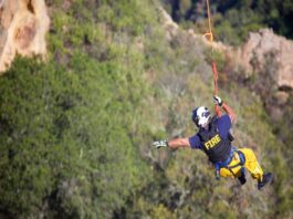 A rescue worker in a shirt labeled “FIRE” hangs off a cliff by a rope. He points towards the rock face of the cliff.