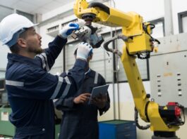 Two men in jumpsuits look at a yellow robotic arm positioned in front of a closed electrical enclosure.