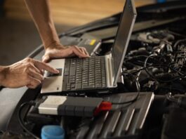 A close-up of a person using a laptop that is sitting inside the open hood of a car and connected to the engine.