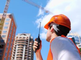 A male construction worker in an orange safety vest and hard hat holding a two-way radio on an outdoor construction site.