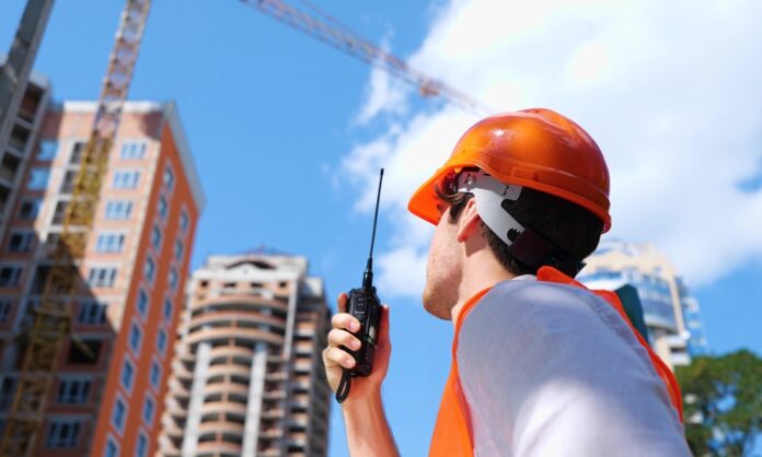 A male construction worker in an orange safety vest and hard hat holding a two-way radio on an outdoor construction site.