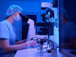 A lab technician wearing scrubs and a hair net is using a micromanipulator in a darkened lab room.