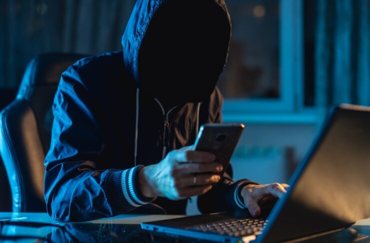 A person wearing black clothing sits in a dark room while typing on a laptop and holding a phone in their hand.