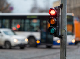 A city traffic light attached to a gray street pole. The red light is illuminated, and vehicles are stopped behind it.