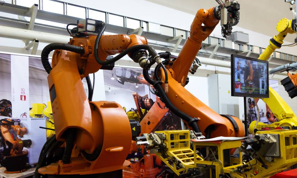 Two large orange robotic arms and a yellow one in a manufacturing facility with white walls and bright lighting.