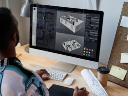 An over-the-shoulder view of a guy working on a gray 3D rendering of a home's floor plan on a monitor at a desk.