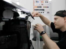 A man carefully handling a vinyl wrap as he applies it to a shiny black car inside a garage workspace.