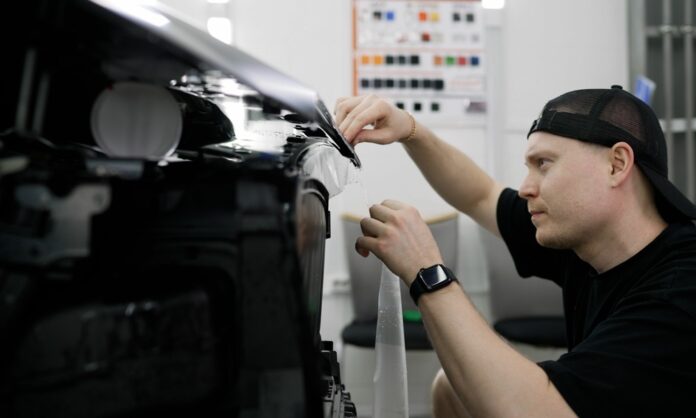 A man carefully handling a vinyl wrap as he applies it to a shiny black car inside a garage workspace.