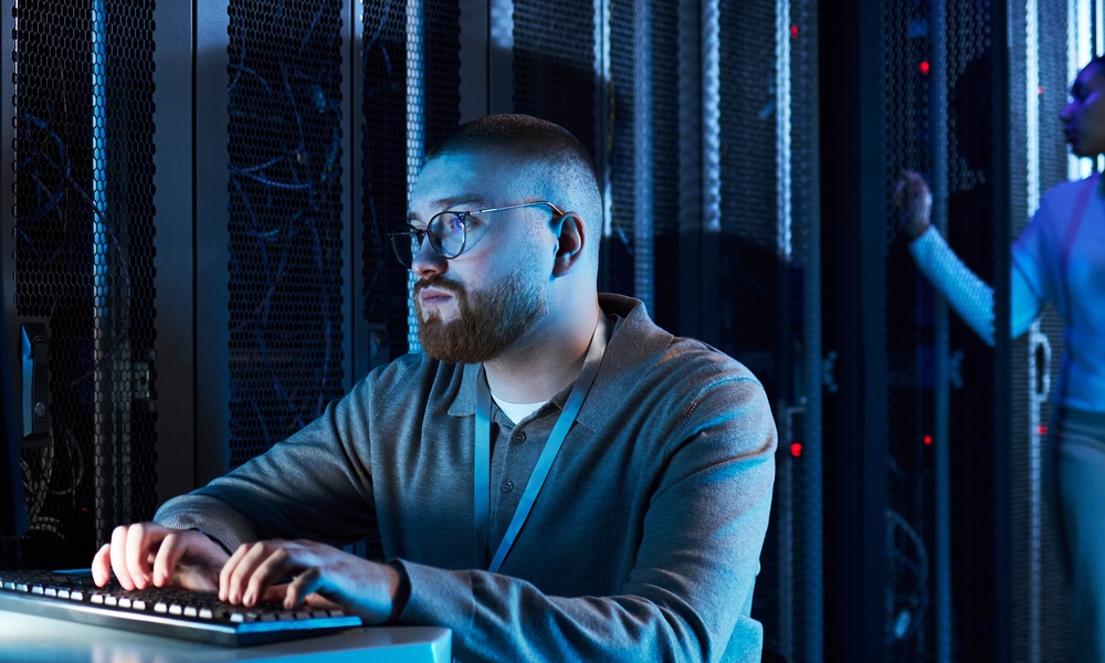 A man in a gray shirt typing on a keyboard while looking at the monitor. A woman checks server room equipment nearby.