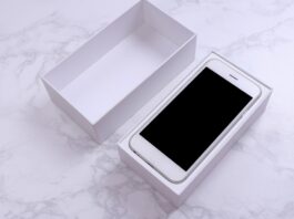 A white smartphone sits in an open white box with a black screen. The lid of the box is next to the bottom on a marble surface.