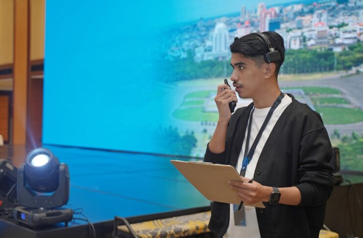 An event organizer stands by a stage set, holding a clipboard and speaking into a two-way radio to coordinate event details.
