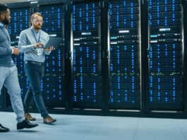 An IT technician carrying a laptop is talking with a Black male engineer colleague as they walk past data server racks.