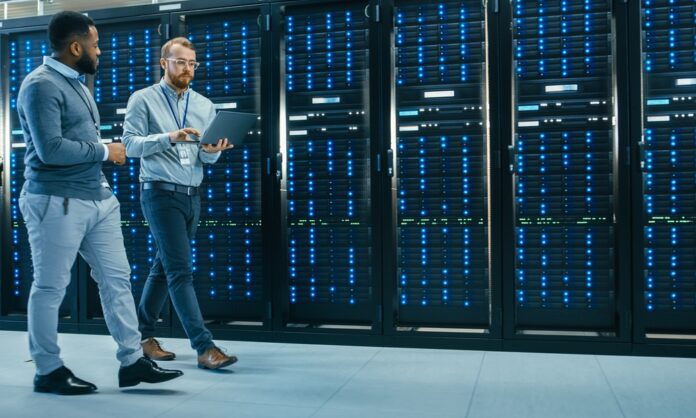 An IT technician carrying a laptop is talking with a Black male engineer colleague as they walk past data server racks.