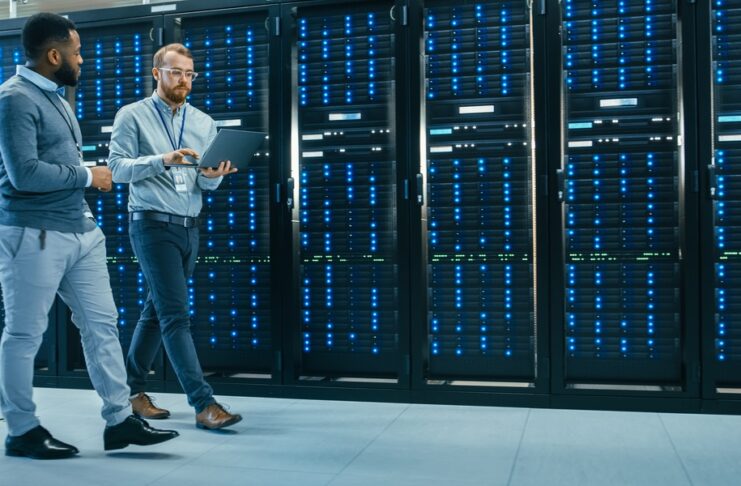 An IT technician carrying a laptop is talking with a Black male engineer colleague as they walk past data server racks.