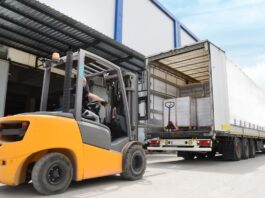 A man operating a yellow forklift drives behind a parked freight shipping truck near a warehouse. The forklift is yellow.