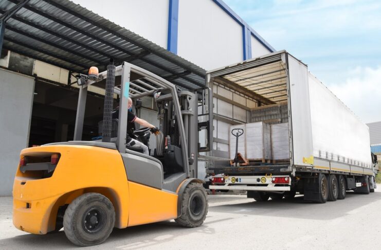 A man operating a yellow forklift drives behind a parked freight shipping truck near a warehouse. The forklift is yellow.