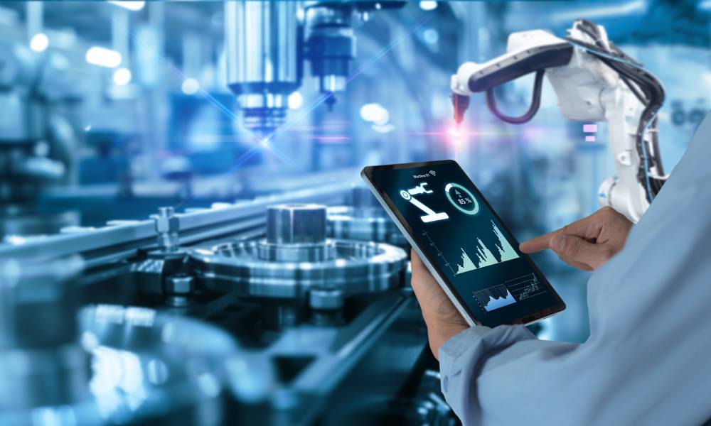 An over-the-shoulder view of an engineer holding a tablet next to a production assembly line with automated robots.