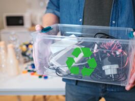 A person holds a container with a green recycling logo, as it's filled with various electronics and accessories.