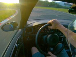 A driver turns the steering wheel with both hands as he turns left on a race track. The sun is on the horizon.