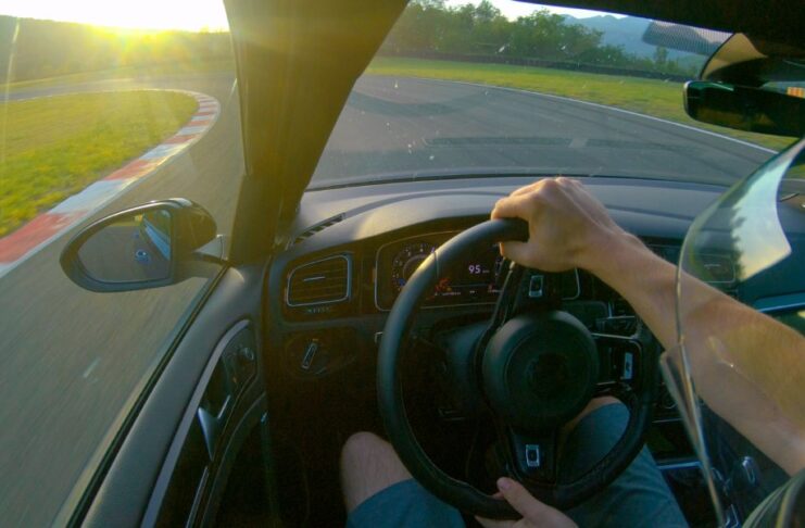 A driver turns the steering wheel with both hands as he turns left on a race track. The sun is on the horizon.