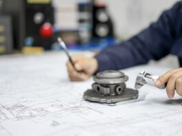 A close-up of a person inspecting a small metal automotive part as they write on a large blueprint at a desk.
